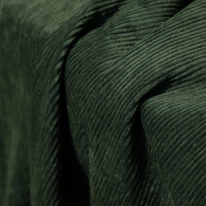 May include: Close-up of a dark green corduroy fabric. The fabric has a ribbed texture with vertical lines and a soft, velvety appearance. The folds and shadows create depth and dimension, highlighting the fabric's texture.