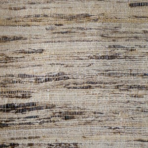 May include: Close-up of a woven rug with a natural, textured appearance. The rug features horizontal bands of light beige and brown fibers, creating a striped pattern. The weave is tight, with visible variations in color and texture.