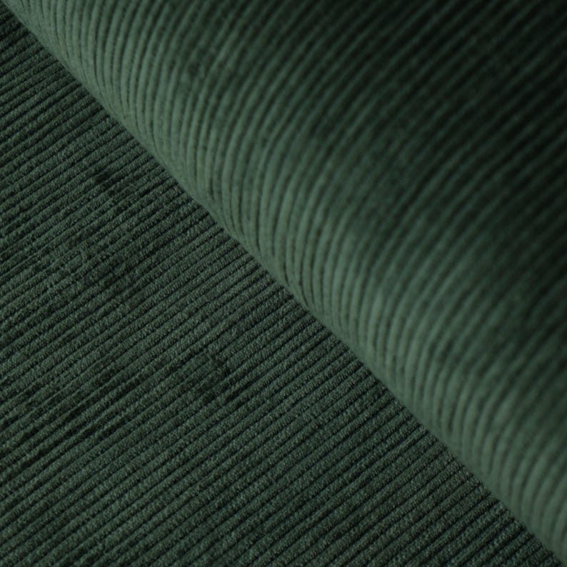 May include: Close-up of a dark green corduroy fabric. The fabric has a ribbed texture with vertical lines. The material is folded over, revealing the texture and color variations. The fabric appears soft and plush.