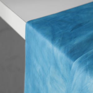 Puede incluir: Primer plano de una tela azul turquesa transparente drapeada sobre la esquina de una mesa blanca. La tela tiene un aspecto suave y texturizado y es ligeramente transl&uacute;cida, creando una est&eacute;tica delicada y aireada. El camino de mesa es ideal para bodas y eventos.