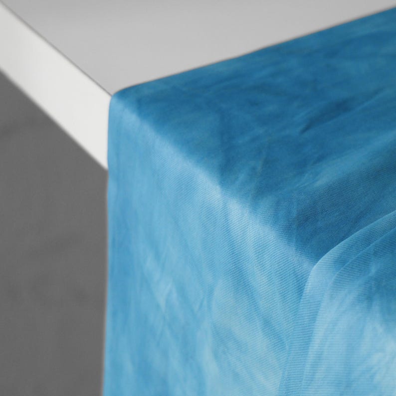 Puede incluir: Primer plano de una tela azul turquesa transparente drapeada sobre la esquina de una mesa blanca. La tela tiene un aspecto suave y texturizado y es ligeramente transl&uacute;cida, creando una est&eacute;tica delicada y aireada. El camino de mesa es ideal para bodas y eventos.