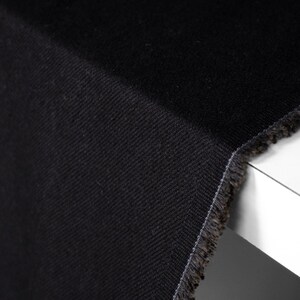 Puede incluir: Primer plano de una tela vaquera negra con un patr&oacute;n de tejido diagonal. La tela tiene un borde sin rematar con hilos deshilachados visibles. El material est&aacute; doblado sobre una superficie blanca.