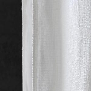 May include: Close-up of white fabric with a textured weave. The fabric is slightly translucent, revealing a glimpse of the dark background. The edge of the fabric is visible, showing a raw, unfinished seam.