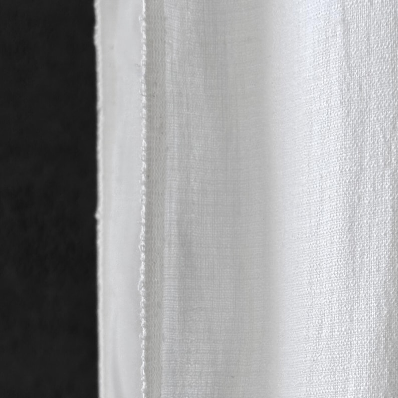 May include: Close-up of white fabric with a textured weave. The fabric is slightly translucent, revealing a glimpse of the dark background. The edge of the fabric is visible, showing a raw, unfinished seam.