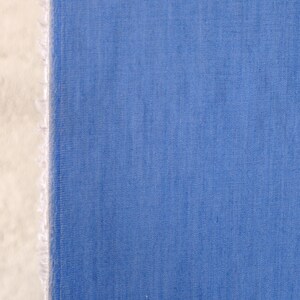 May include: A close-up of blue fabric with a textured weave. The fabric has a raw, unfinished edge on the left, showing a white, frayed texture. The blue is a solid colour, evenly distributed across the material.