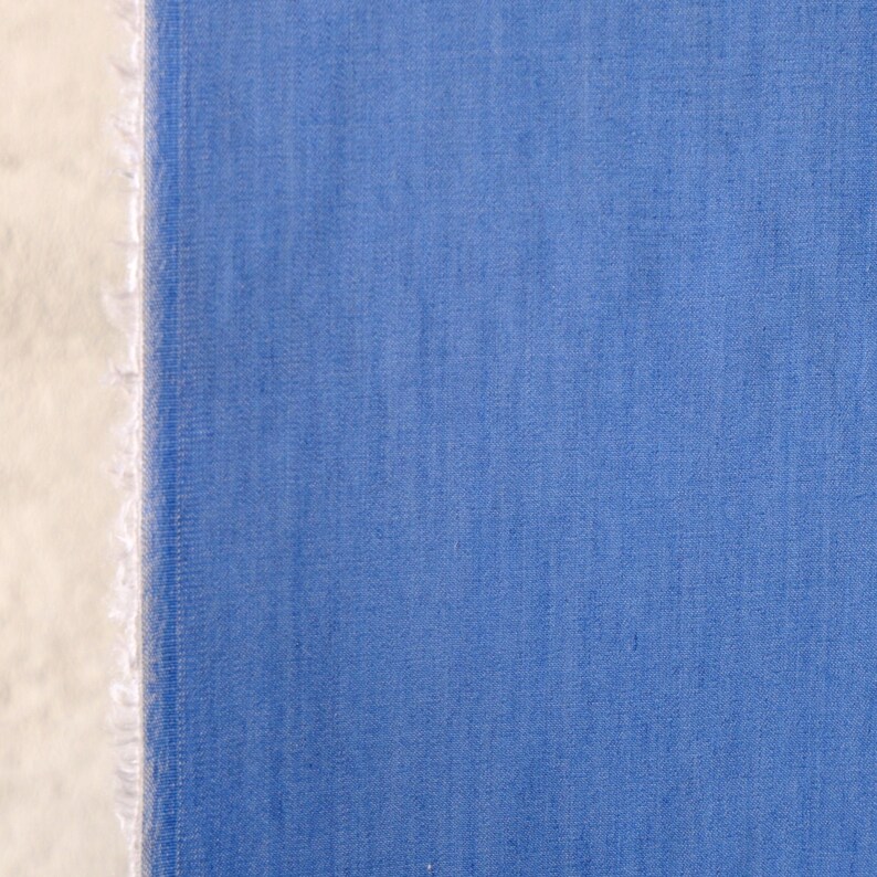 May include: A close-up of blue fabric with a textured weave. The fabric has a raw, unfinished edge on the left, showing a white, frayed texture. The blue is a solid colour, evenly distributed across the material.