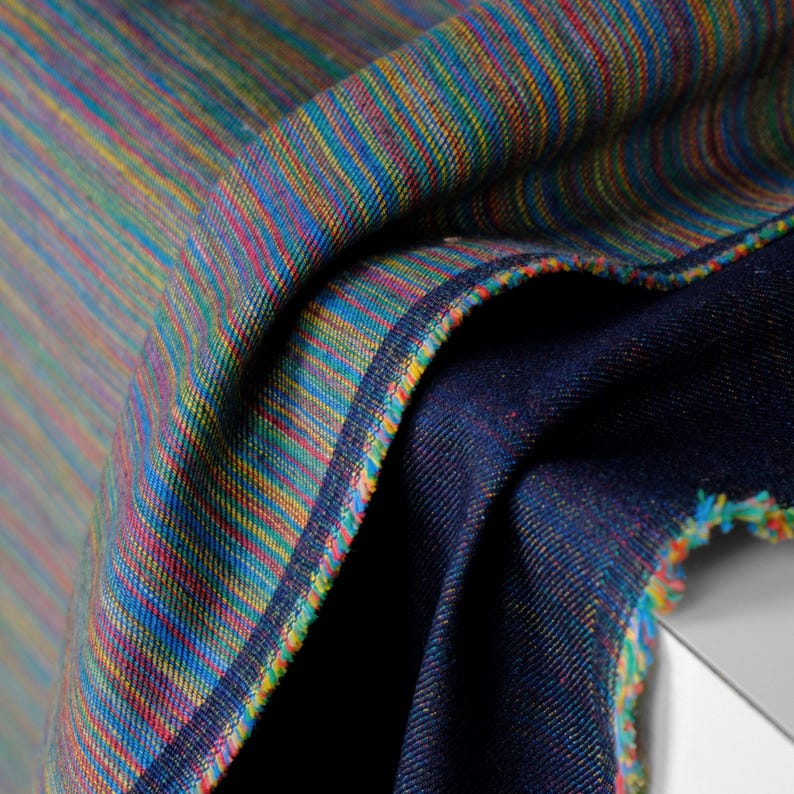 Op de afbeelding: Close-up van een gevouwen stof met een levendig gestreept patroon. De stof heeft een mix van kleuren, waaronder blauw, groen, geel en rood. De randen zijn afgewerkt met een contrasterende donkerblauwe stof en kleurrijke stiksels.