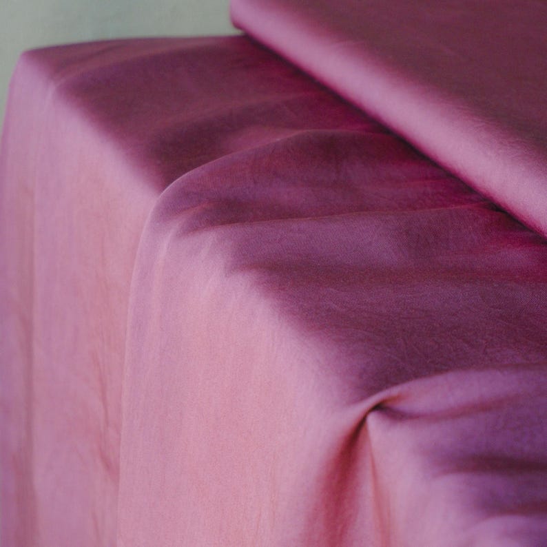 Pode incluir: Um close-up de tecido drapeado em tons de rosa e roxo. O tecido tem uma textura macia e fluida com varia&ccedil;&otilde;es sutis de cor, criando um efeito de gradiente. As dobras e sombras adicionam profundidade e dimens&atilde;o ao material.