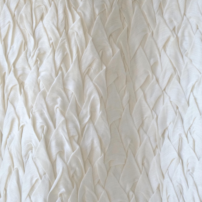 May include: Off-white fabric with a textured, layered design. The fabric is arranged in a repeating pattern of folded, triangular shapes, creating a sense of depth and dimension. The overall effect is soft and visually interesting.