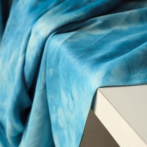 Puede incluir: Primer plano de una tela azul te&ntilde;ida con efecto tie-dye sobre una superficie blanca. La tela tiene un efecto acuarela con tonos de azul claro y oscuro. La tela es suave y fluida, con textura y pliegues visibles.