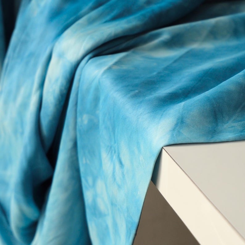 Puede incluir: Primer plano de una tela azul te&ntilde;ida con efecto tie-dye sobre una superficie blanca. La tela tiene un efecto acuarela con tonos de azul claro y oscuro. La tela es suave y fluida, con textura y pliegues visibles.