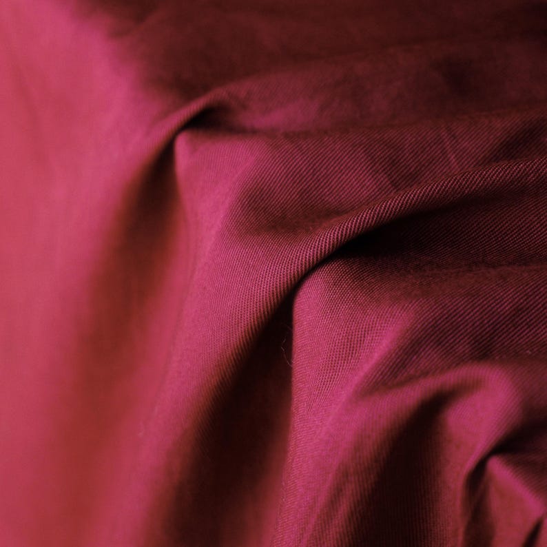 Pode incluir: Close-up de um tecido magenta escuro com tecido diagonal vis&iacute;vel. O tecido &eacute; dobrado e drapeado, criando sombras e destaques que enfatizam sua textura e cor. O material parece ser um algod&atilde;o de peso m&eacute;dio ou um t&ecirc;xtil semelhante.