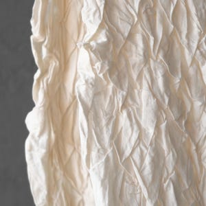 May include: Close-up of a textured, off-white fabric. The fabric has a quilted pattern with diamond shapes. The material appears to be soft and slightly wrinkled, with shadows adding depth to the design. The background is a solid gray.