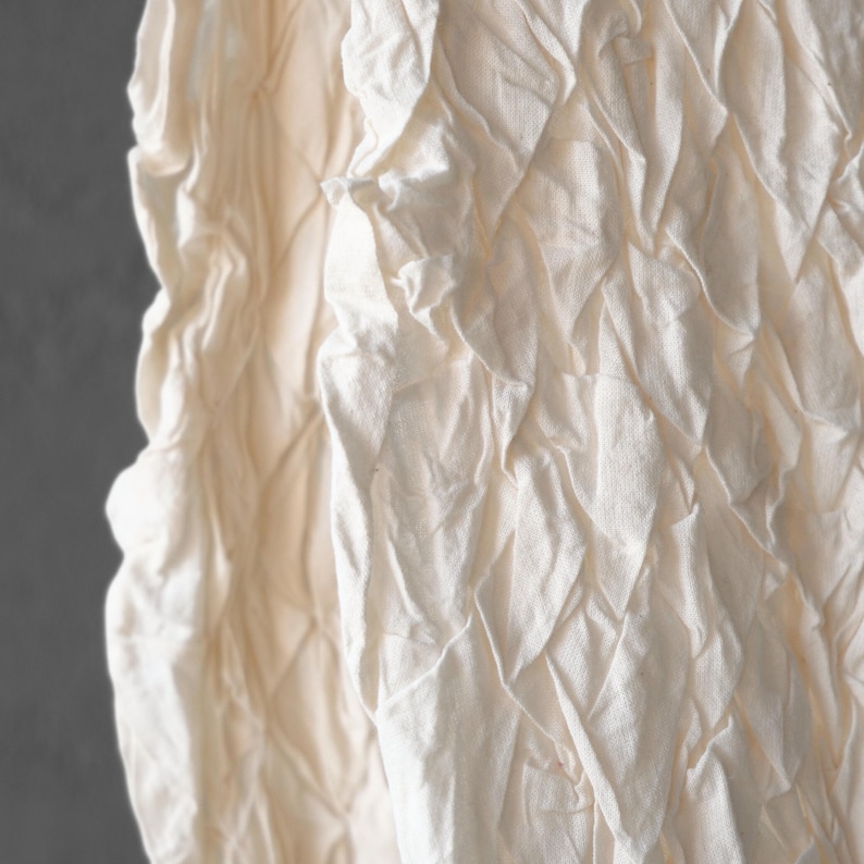 May include: Close-up of a textured, off-white fabric. The fabric has a quilted pattern with diamond shapes. The material appears to be soft and slightly wrinkled, with shadows adding depth to the design. The background is a solid gray.