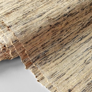 May include: Close-up of a rolled-up, textured rug. The rug is woven with natural fibers in shades of beige and brown, with dark brown stripes running across the surface. The edges of the rug are visible, showing the layered texture.