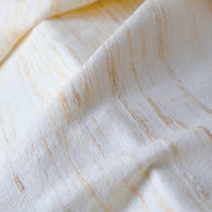 May include: Close-up of a folded, off-white fabric with vertical, light-brown stripes. The fabric has a textured appearance, with visible woven threads and subtle variations in color. The folds create shadows and depth, highlighting the material's softness.