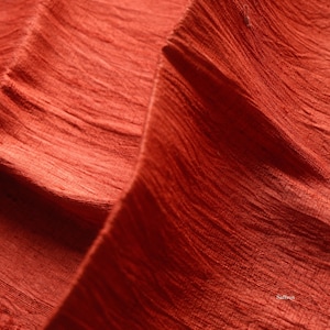 May include: Close-up of a textured, rust-colored fabric with visible folds and a subtle sheen. The fabric's surface has a ribbed pattern, creating depth and visual interest. The overall impression is of a soft, flowing material.
