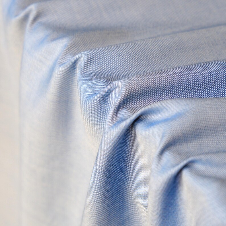 May include: Close-up of light blue fabric with visible texture and folds. The fabric has a subtle sheen and a gradient effect, transitioning from a lighter shade to a deeper blue. The image highlights the material's drape and texture.