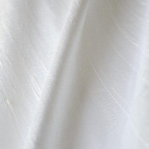 May include: Close-up of white fabric with subtle vertical and diagonal textures. The fabric is folded, creating shadows and highlights that emphasize the texture and drape of the material. The overall appearance is clean and minimalist.