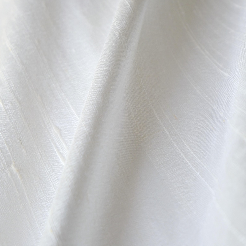 May include: Close-up of white fabric with subtle vertical and diagonal textures. The fabric is folded, creating shadows and highlights that emphasize the texture and drape of the material. The overall appearance is clean and minimalist.