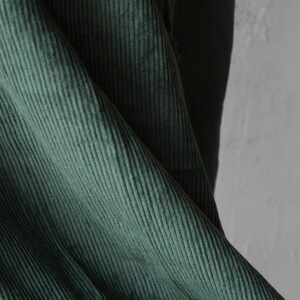 May include: Close-up of a section of dark green corduroy fabric. The fabric has a ribbed texture with vertical lines. The folds and shadows create depth and dimension. The corduroy appears soft and plush.