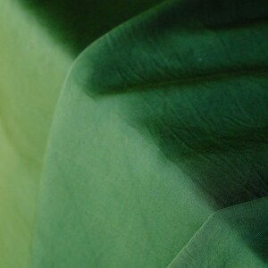 Pode incluir: Close-up de um tecido dobrado em tons de verde. O tecido apresenta um gradiente, passando de um verde claro, quase amarelo-esverdeado, a um verde floresta profundo. A textura &eacute; vis&iacute;vel, com dobras e sombras sutis.