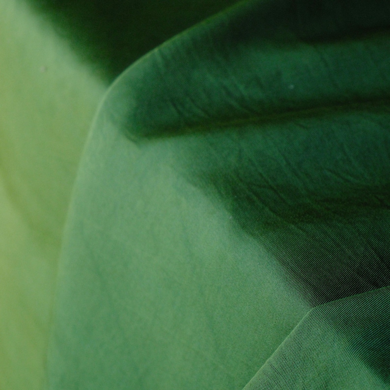 Pode incluir: Close-up de um tecido dobrado em tons de verde. O tecido apresenta um gradiente, passando de um verde claro, quase amarelo-esverdeado, a um verde floresta profundo. A textura &eacute; vis&iacute;vel, com dobras e sombras sutis.