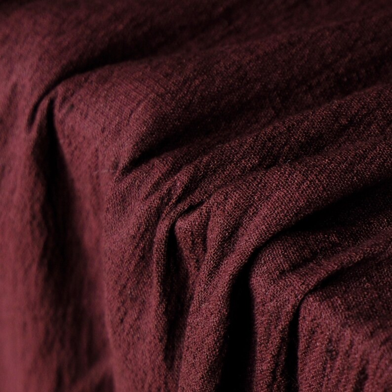 May include: Close-up of a textured, burgundy fabric with visible woven threads. The fabric is draped, creating soft folds and shadows. The deep red color suggests a luxurious textile, possibly linen or a similar material.