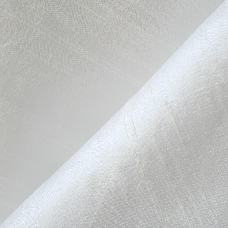 May include: Close-up of white fabric with a subtle, textured weave. The fabric is folded diagonally, revealing the texture and sheen of the material. The surface has a slight sheen, suggesting a luxurious or high-quality textile.