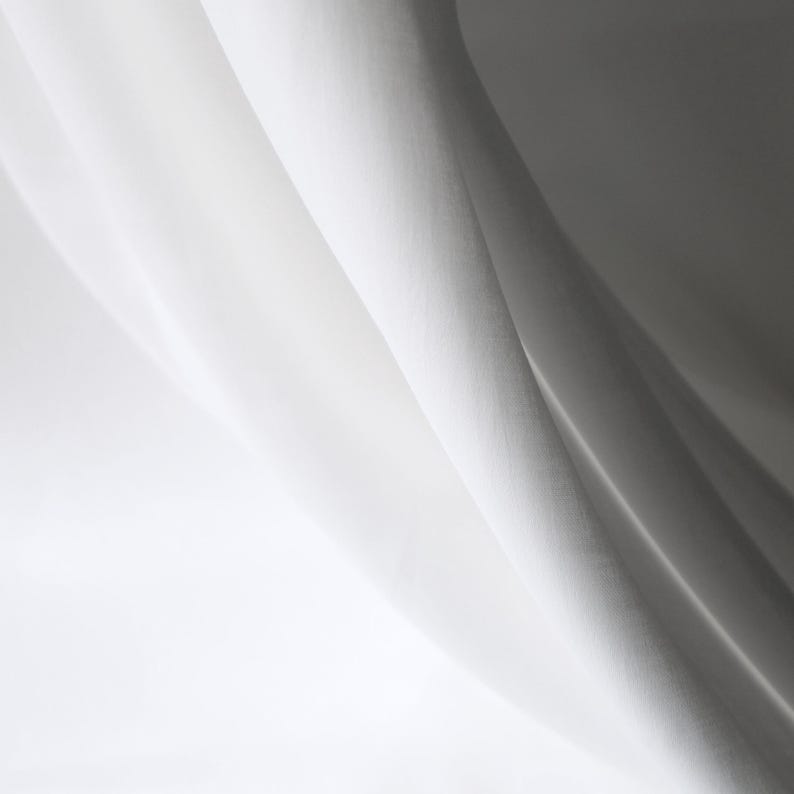 Puede incluir: Primer plano de tela blanca con pliegues suaves y sombras. La tela parece ligera, posiblemente lino o similar, con una textura sutil. La iluminaci&oacute;n crea un efecto de gradiente, pasando de blanco brillante a un gris m&aacute;s oscuro.