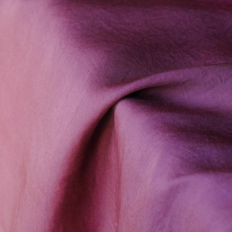 Pode incluir: Close-up de um tecido dobrado em tons de roxo e rosa. O tecido tem um brilho sutil e textura vis&iacute;vel, com linhas diagonais que adicionam profundidade. As dobras criam sombras e destaques.