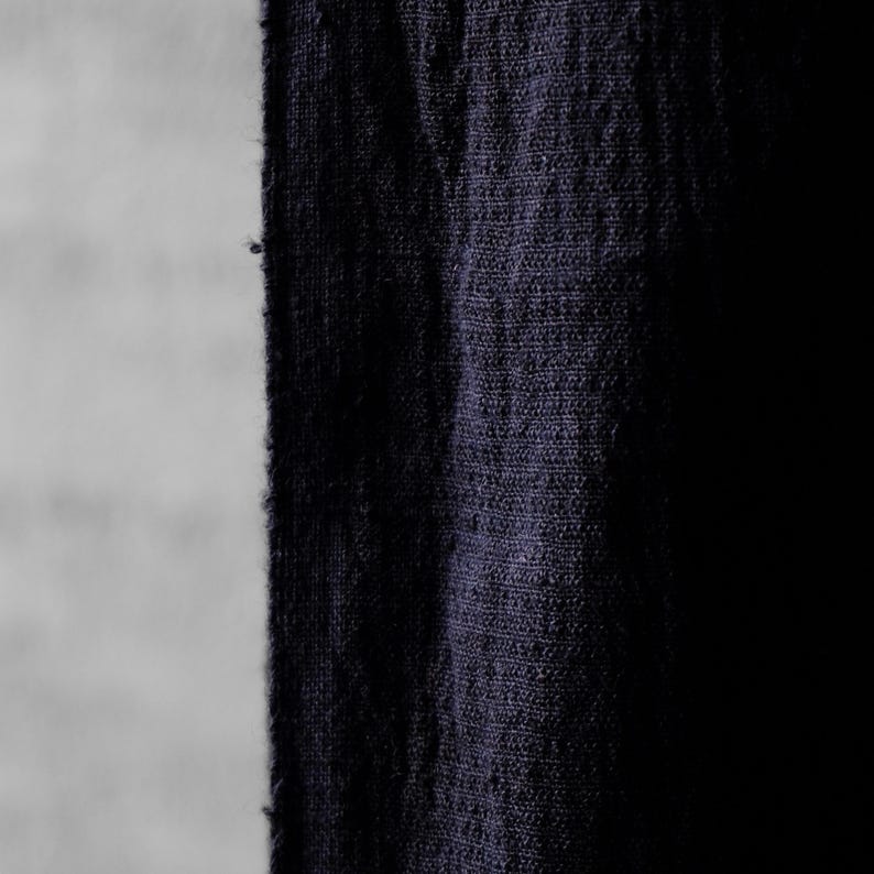 May include: Close-up of a dark blue fabric with a textured weave. The fabric appears to be a heavy linen or similar material, with a slightly frayed edge. The background is a light gray, providing contrast.