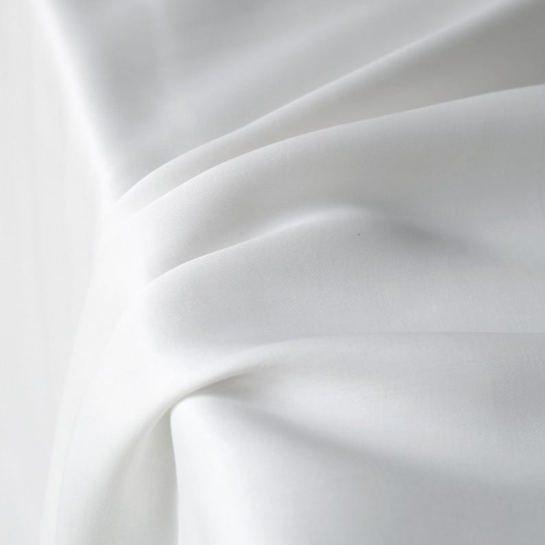 Puede incluir: Primer plano de tela blanca con pliegues suaves y sombras sutiles. La tela parece suave y fluida, creando una sensaci&oacute;n de movimiento y textura. La composici&oacute;n general es minimalista y elegante.