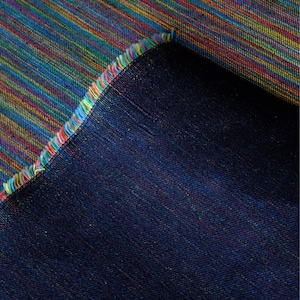 Op de afbeelding: Close-up van een textiel met een levendig, veelkleurig streepjespatroon. De stof heeft een mix van blauwe, gele, rode en groene tinten. De rand van de stof is gefranjerd met een regenboog aan kleuren, en de achterkant is effen donkerblauw.