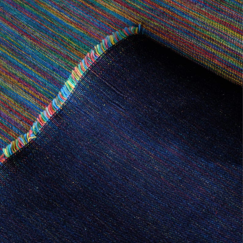 Op de afbeelding: Close-up van een textiel met een levendig, veelkleurig streepjespatroon. De stof heeft een mix van blauwe, gele, rode en groene tinten. De rand van de stof is gefranjerd met een regenboog aan kleuren, en de achterkant is effen donkerblauw.