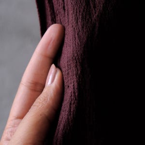 May include: Close-up of a textured, dark burgundy fabric. The fabric is held by a hand with a light pink nail polish. The background is a gradient of gray and black.