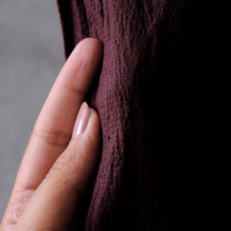 May include: Close-up of a textured, dark burgundy fabric. The fabric is held by a hand with a light pink nail polish. The background is a gradient of gray and black.
