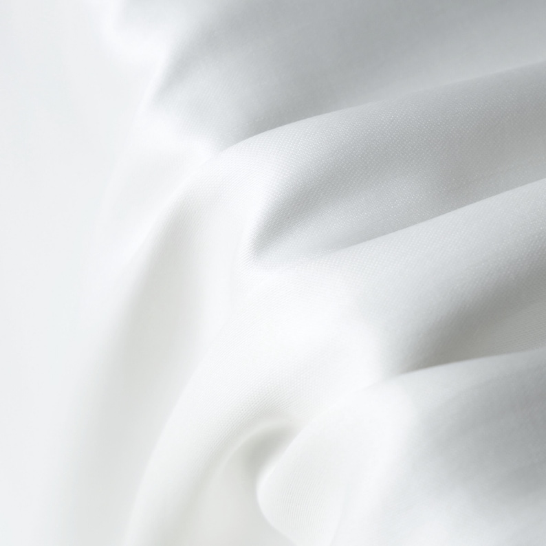 Puede incluir: Primer plano de tela blanca con pliegues suaves y sombras sutiles. La tela parece lisa y fluida, con un ligero brillo. La imagen resalta la textura y la ca&iacute;da del material, sugiriendo un textil lujoso.