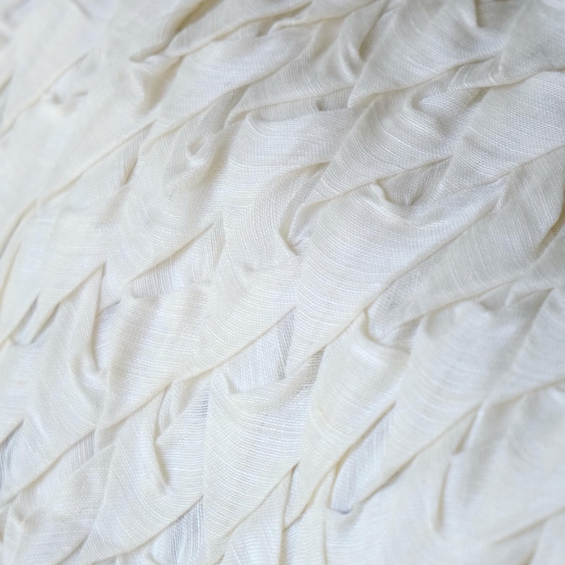 May include: Close-up of a white fabric with a textured, layered design. The fabric appears to be soft and has a rippled effect, creating a pattern of folds and ridges. The color is a bright, clean white.