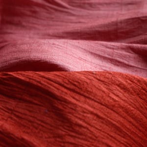 May include: Close-up of three layers of fabric in shades of red. The top layer is a muted rose, the middle layer is a lighter pink, and the bottom layer is a deep, textured red. The fabric appears soft and flowing.