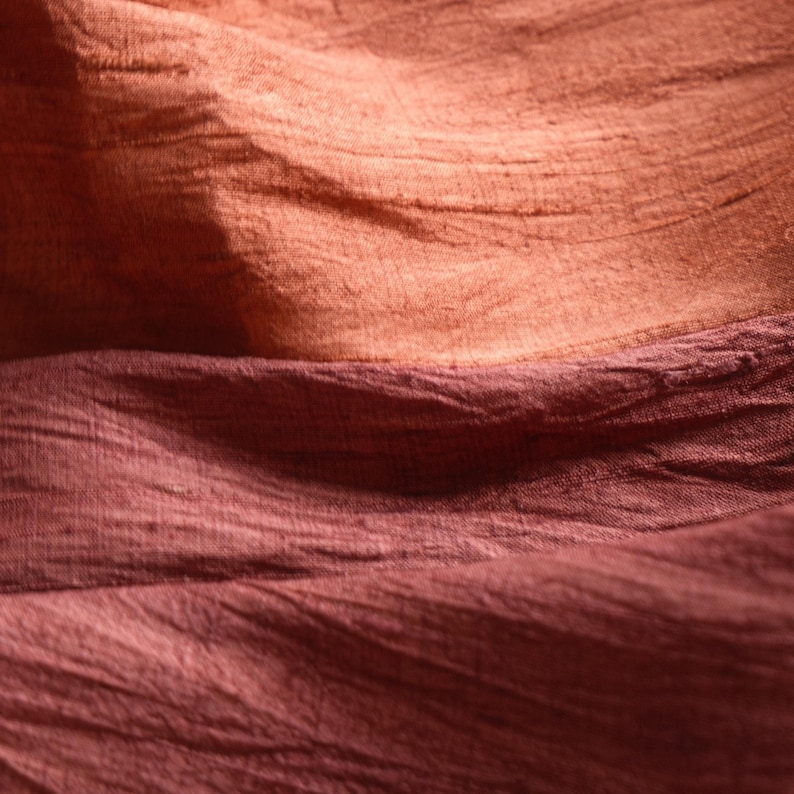 May include: Close-up of textured fabric in shades of peach and burgundy. The fabric appears to be layered, with the peach color on top and the burgundy below. The fabric has a slightly wrinkled appearance, suggesting it may be silk or a similar material.