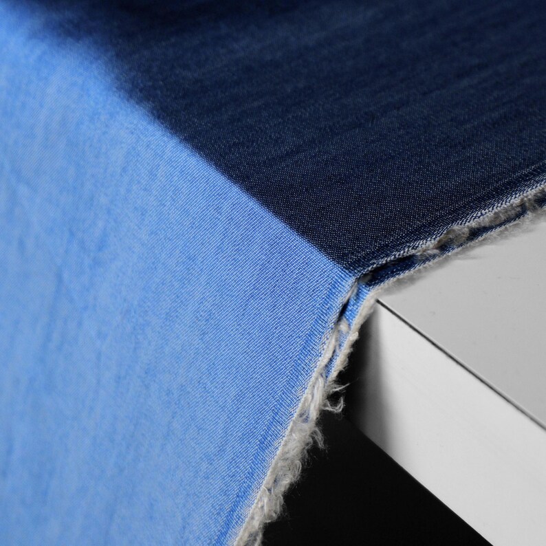 May include: A close-up shot of denim fabric in two shades of blue. The fabric is split diagonally, with a lighter blue on the left and a darker blue on the right. The edges are frayed, revealing the raw texture of the material.