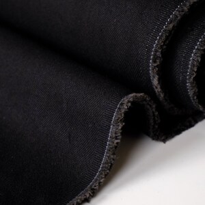 Puede incluir: Primer plano de tela vaquera negra, tejida apretadamente con un patr&oacute;n de sarga diagonal. Los bordes est&aacute;n deshilachados, revelando la textura de la tela. El color oscuro y la textura sugieren que es adecuada para varias prendas.