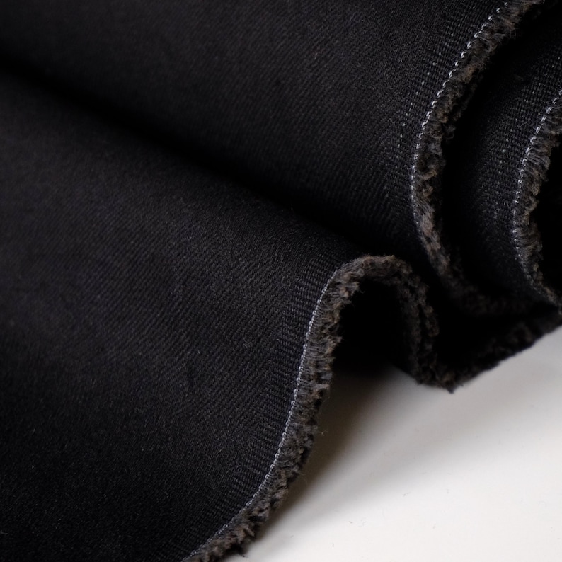 Puede incluir: Primer plano de tela vaquera negra, tejida apretadamente con un patr&oacute;n de sarga diagonal. Los bordes est&aacute;n deshilachados, revelando la textura de la tela. El color oscuro y la textura sugieren que es adecuada para varias prendas.