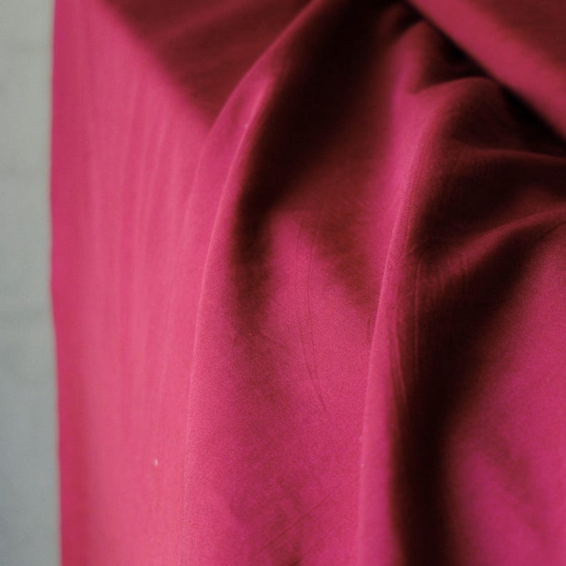 Pode incluir: Close-up de um tecido magenta profundo, com dobras e sombras suaves. O material parece liso e fluido, sugerindo um tecido leve. A cor &eacute; rica e saturada, preenchendo a moldura.