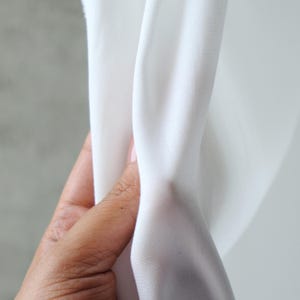 Pode incluir: Close-up de tecido branco segurado por uma m&atilde;o. O tecido &eacute; liso e parece ser um material leve, possivelmente algod&atilde;o ou uma mistura. O tecido est&aacute; drapeado, mostrando dobras e textura.