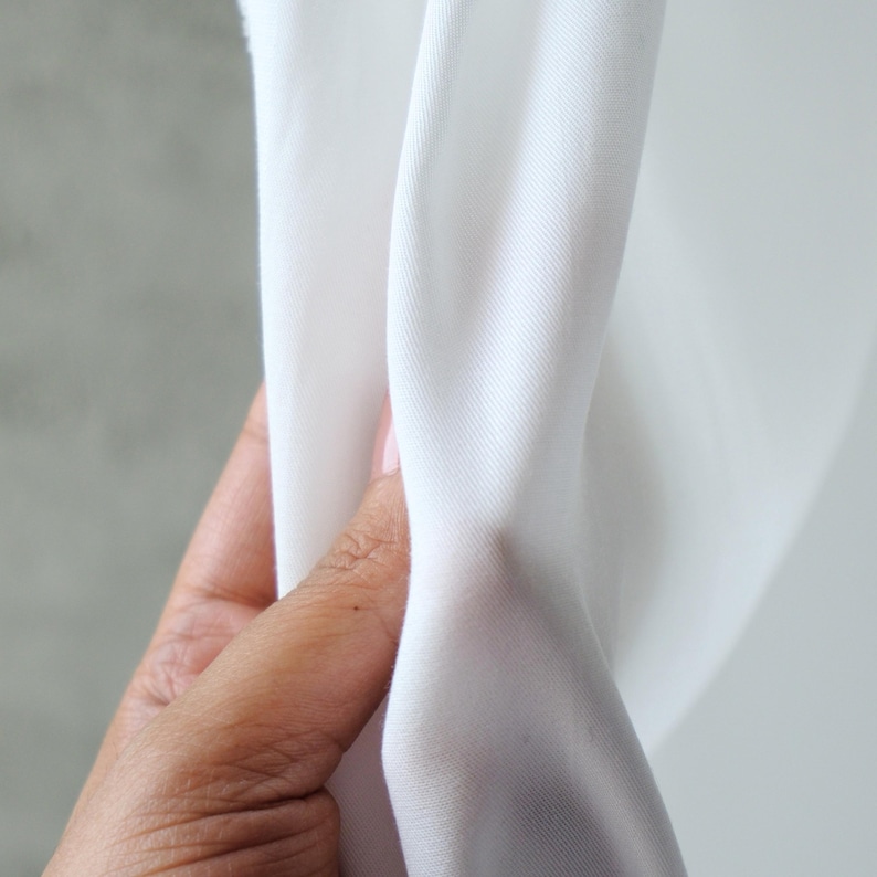 Pode incluir: Close-up de tecido branco segurado por uma m&atilde;o. O tecido &eacute; liso e parece ser um material leve, possivelmente algod&atilde;o ou uma mistura. O tecido est&aacute; drapeado, mostrando dobras e textura.