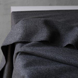 May include: A close-up of a dark gray fabric with a soft, textured appearance. The fabric is draped over a white surface, creating folds and shadows. The material appears to be a heavy weight, possibly wool or a wool blend.