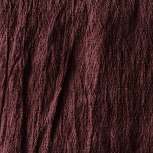 May include: Close-up of a textured, burgundy fabric. The material appears to be slightly wrinkled, with visible folds and variations in the weave. The color is a deep, rich shade of red-purple.