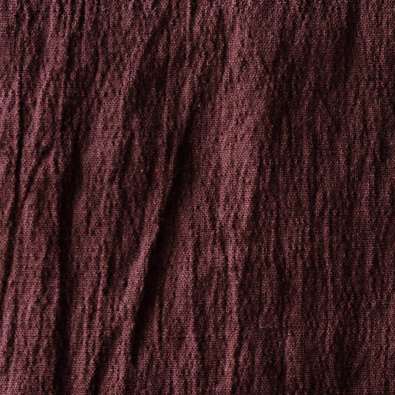 May include: Close-up of a textured, burgundy fabric. The material appears to be slightly wrinkled, with visible folds and variations in the weave. The color is a deep, rich shade of red-purple.